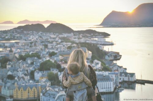 maman avec un bébé en porte-bébé devant un paysage d'un bord de mer avec soleil couchant